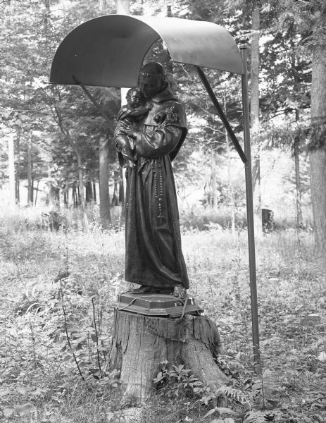 View of a statue of Saint Joseph and a child, covered by a metal shelter. The base of the statue is strapped to a tree stump.