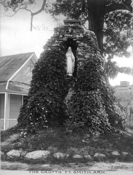 View of the Holy Grotto on the Convent Grounds of the Sisters of Mercy. The grotto is composed of leftover brick and the chimney of the 1857 church building. The Madonna statue stands in a niche in the ivy-covered structure. Caption reads: "The Grotto. Ft. Smith, ARK."  