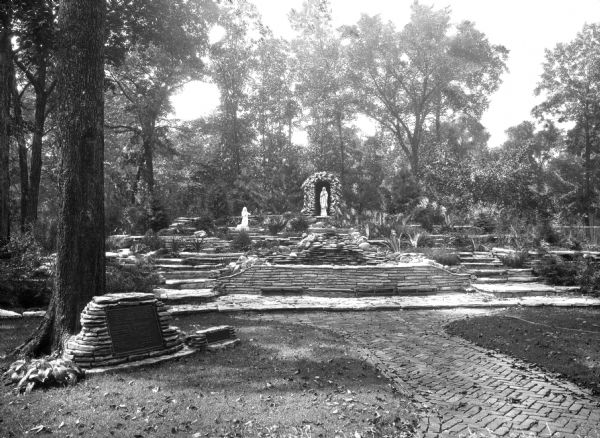 View of the Shrine of Our Lady, a grotto at Rosary College, founded in 1922. A path leads to stone stairs that reach a statue of Mary, as well as a statue of a kneeling figure. On the left, a plaque built into a stone structure marks the grotto.