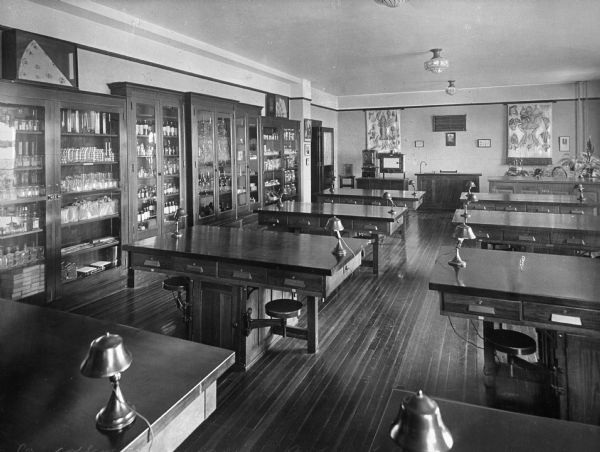 View of a biology laboratory at College Misericordia, established by the Sisters of Mercy on August 15, 1924. There are two rows of tables with lamps, and shelves with glass doors line the wall on the left.