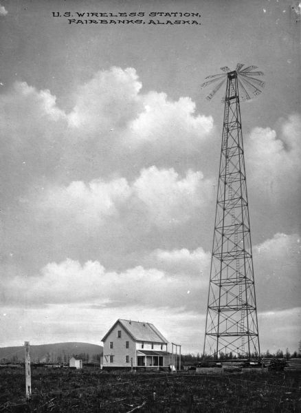 View of a United States Wireless Station. A residential building stands to the left of a tall tower where piles of lumber can be seen. Caption reads: "U.S. Wireless Station, Fairbanks, Alaska."