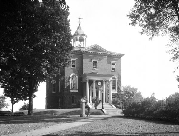 View of Hathorn Hall at Bates College. Completed in 1857, the hall is a small brick building featuring a bell tower. A man is seen walking on the sidewalk to the left of the steps.