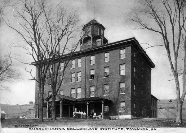 View of the Susquehanna Collegiate Institute, founded in 1858 by the Evangelical Lutheran Church as the Missionary Institute and Susquehanna Female College. A group of men and women pose outside the building, and at a third floor window, a man can be seen looking outward. Caption reads: "Susquehanna Collegiate Institute, Towanda, PA."