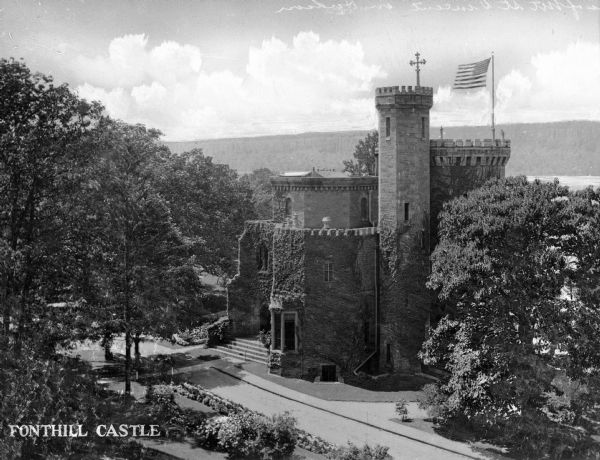 Elevated view of Fonthill Castle, built in 1852. The ivy-covered castle began as the convent for the College of Mount Saint Vincent, founded by the Sisters of Charity of New York in 1847. The structure, topped with a cross and United States flag, overlooks the Hudson River. Caption reads: "Fonthill Castle."