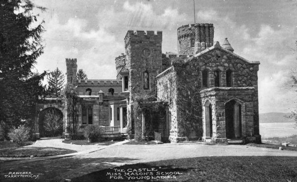 Exterior of Miss Mason's School for Young Ladies, designed by A. J. Davis and completed in 1856. The stone building features a porte cochère, sheltered entries with arches and castle towers. The Hudson River is in the background on the right. Caption reads: "'The Castle' Miss Mason's School For Young Ladies."