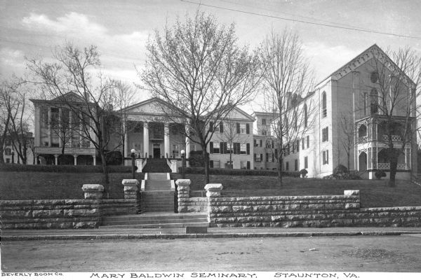 View from across a road of a row of buildings of Mary Baldwin Seminary, established in 1842. A stone fence and stairs separate the lawn from the sidewalk. Caption reads: "Mary Baldwin Seminary, Staunton, VA."