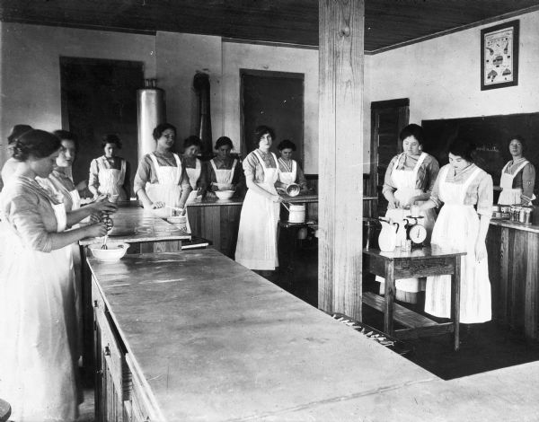 View from across a room of a home economics class at Martha Berry Girls' School, established in 1909. Women gather around an area enclosed by tables and observe a demonstration.