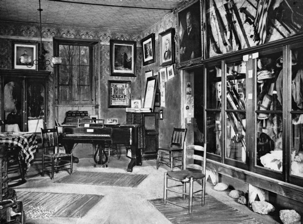 View of a room filled with portraits and relics from the Civil War. A piano is placed below a window across the room, and the walls are lined with framed portraits, paintings, and news articles. On the right, a cabinet holds military uniforms and weapons.