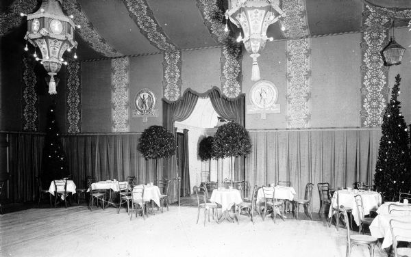 Interior of a nightclub. The room features tables and chairs and is decorated with elaborate drapery, lighting and potted trees.