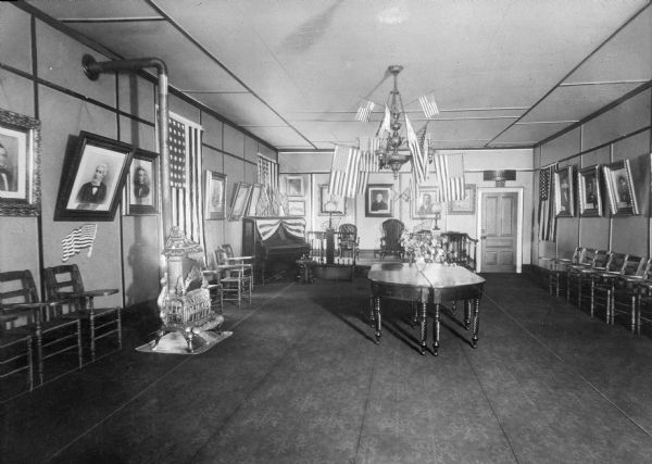 Interior of Memorial Hall at Jefferson College, chartered in 1802. The walls are lined with framed portraits hanging from picture rails, with chairs below. On the left is a furnace and piano, and a table stands in the middle of the room. A chandelier is adorned with United States flags.