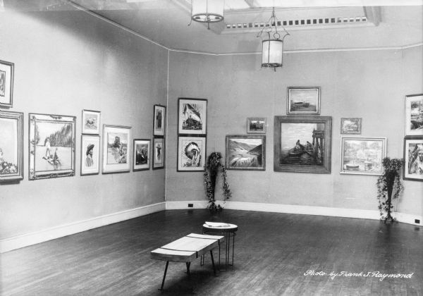 Interior of an art museum. The view includes a bench and table in the middle of the room and walls lined with paintings. Potted plants stand on either side of pieces of art.