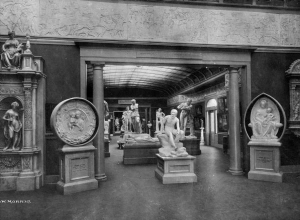 Interior of Parrish Art Museum, built in 1898. The gallery features  sculptures a a bas relief along the ceiling. The room leads to another room through an opening framed by columns.