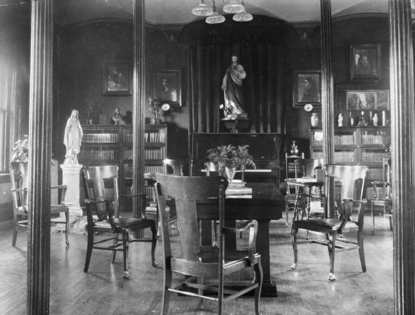 Interior of a small library at Saint Joseph's Academy of Maine, opened in 1912. Chairs are gathered around a table near bookcases. In the room are religious statues, portraits, and paintings.