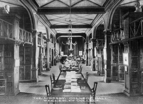 Interior of Wellesley College Library, founded in 1870. The view includes long study tables, and paintings and busts are displayed throughout the room. Bookshelves line the side aisles. Caption reads: "The Library, College Hall, Wellesley College."