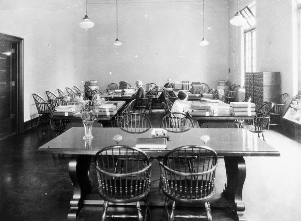 Interior of a library reading room. A man and two women sit at tables, reading. A door leads out of the room on the left, and windows have open transoms on the right.