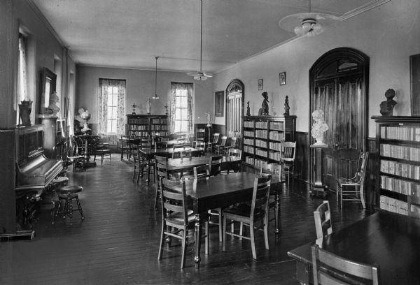 Interior of the library reading room at Holy Angels School, established in 1924. A piano stands against the left wall, and busts, figurines, and bookshelves surround the room, with tables and chairs in the center.