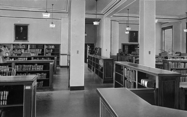Interior of the reading room at Adelphi College, founded in 1863. Rows of bookshelves fill the room, and two portraits hang on the walls.