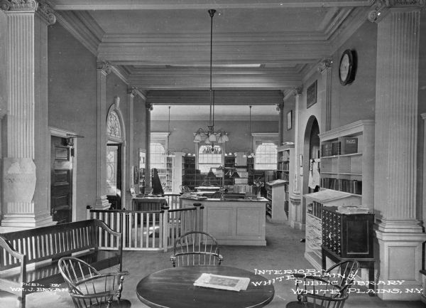 Interior of White Plains Library, chartered in 1899. A chandelier hangs above the reference desk, and bookshelves surround the area. Caption reads: "Interior White Plains Public Library, White Plains, N.Y."