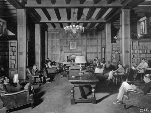 Interior of a reading room at Willard Straight Hall, opened in 1925.  The hall can be found in the union of Cornell University, founded in 1865. Men sit and read in chairs, surrounded by bookshelves. A chandelier hangs above tables and lamps.