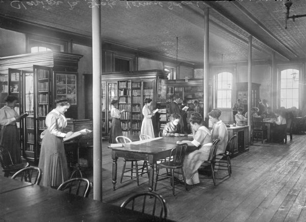 Interior of a library at Clarion State Normal School, established in 1873. Men and women read at tables and choose books from shelves.