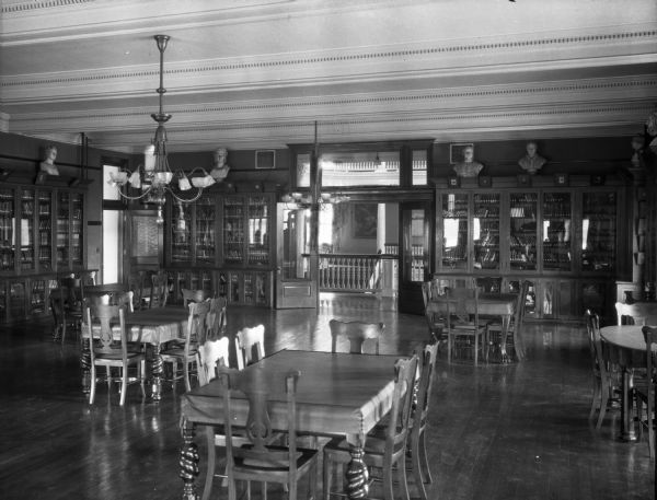 Interior of a library in Saint Joseph Hall, opened in 1903. Located on the campus of Mount Saint Joseph Collegiate Institute, founded in 1900, the library features bookshelves that line the walls of the room and busts that are placed on top of them. Tables and chairs stand in the middle of the room. A set of double doors opens to reveal a five-story rotunda.