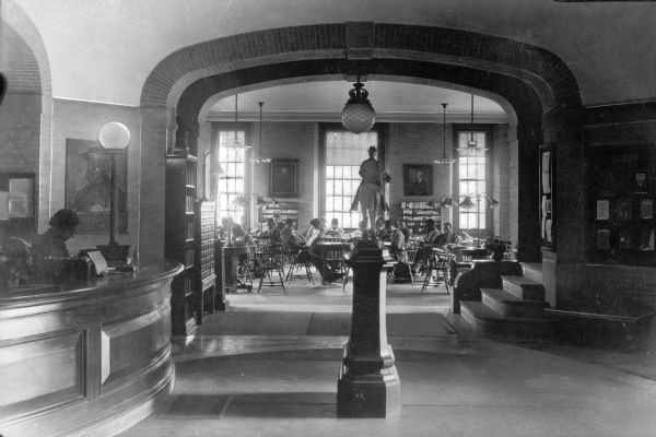Interior of a small reading room at Collis P. Huntington Memorial Library, built in 1903. A woman sits at a reference desk on the left, and other people read at tables in the room beyond an equestrian statue.
