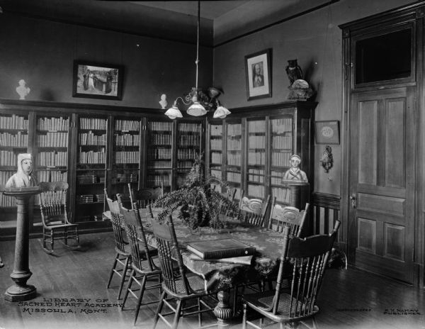 Interior of the library and reading room of Loyola Sacred Heart Academy, established in 1873.  The room features bookshelves along the wall and sculptures.  A table and chairs can be seen in the foreground.  Published by R.H. McKay.