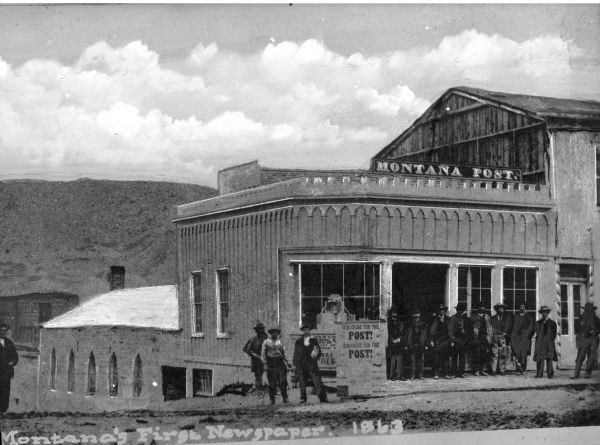 Exterior of the first office of the Montana Post Newspaper, the first newspaper in Montana, built in 1863.  Men stand in front of the building near a sign that reads, "Subscribe for the Post!"