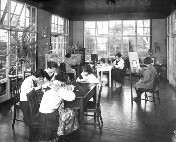 Interior of an art room. Female students are sitting at tables and easels, painting or doing craft projects. Large windows open to a garden outside.