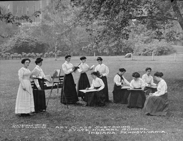 View of woman at an outdoor art class on a lawn, sketching at State Normal School, which opened in 1875. The group of women are standing and sitting outdoors drawing and painting. There is a school building in the background among trees. Caption reads: "Art Class Sketching, State Normal School, Indiana, Pennsylvania."