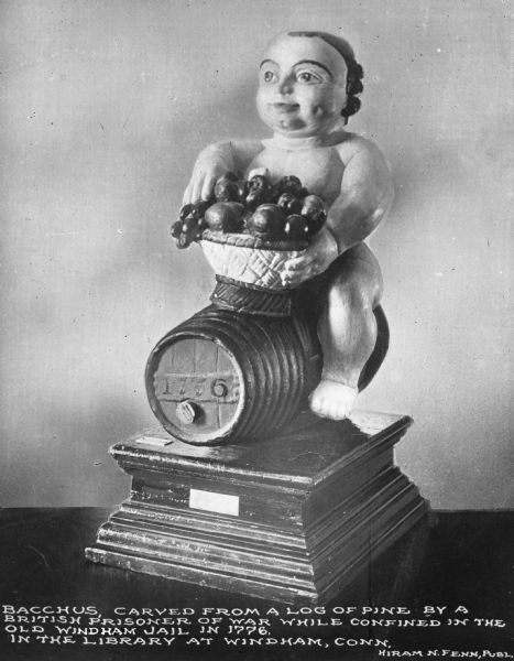 View of a carved, wooden figurine of Bacchus, God of Wine. The sculpture is located in the city's library. Caption reads: "Carved from a log of pine by a British prisoner of war while confined in the Old Windham Jail in 1776. In the library at Windham, Conn."