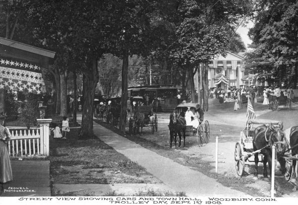 View from lawn toward the street where a trolley runs, and horse-drawn vehicles parked along the curbs. People stand on the lawns and on porches, facing toward Old Town Hall, built in 1846. Caption reads: "View showing cars and town hall, Woodbury, Conn. Trolley Day, September 1, 1908."