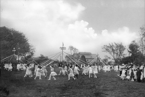 View of children swinging around maypoles on May Day at Hampton Institute, established 1868. Campus buildings are in the background.