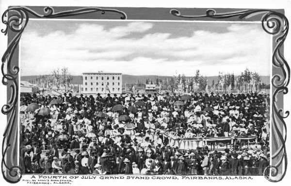 Elevated view of a crowd in a grandstand on the Fourth of July. The grandstand is decorated with flags and banners. Buildings of Fairbanks are in the background. Caption reads: "A Fourth of July Grand Stand Crowd, Fairbanks, Alaska."