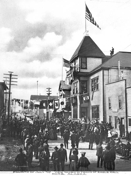 Elevated view of a Fourth of July street scene taken in front of Eagle's Hall. The Hall was built by the Fraternal Order of Eagles, a group founded in 1898. Crowds gather in the street and flags adorn the rooftops. A banner of flags is hanging above the street. Caption reads: "Fourth of July '07. View taken in front of Eagle Hall. Nome, Alaska."