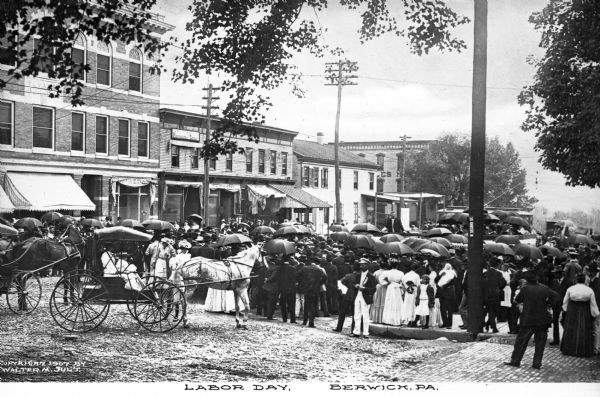 View of a crowd on Labor Day. The street scene is on W. Second Street where horse-drawn vehicles are parked. To the left of the crowd is the Evans Building, built in 1905n. To its right stands a photography studio and an awning labeled W. C. McKinney. Caption reads: "Labor Day, Berwick, PA."