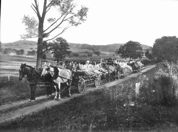 Slightly elevated view of a hayride at Camp Sherwood. Children and adults are sitting on hay in horse-drawn vehicles. A field and hills are in the background.
