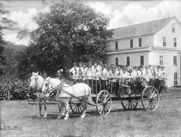 View of a hayride on the lawn outside a residential-type building. A group of girls stand in a wagon driven by two horses. Banners line the side of the wagon that reads: "HNC."