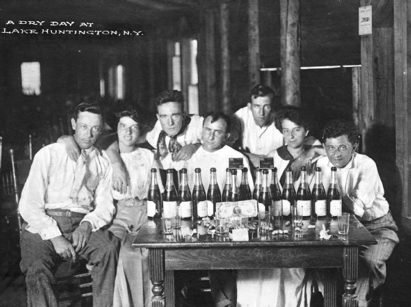 A group of people sitting at a table and posing near two packs of Pall Mall cigarettes balanced on empty bottles. Empty glasses, peanut shells, and corks fill the table where a one hundred dollar bill is propped up between glasses. Caption reads: "A Dry Day at Lake Huntington, NY."