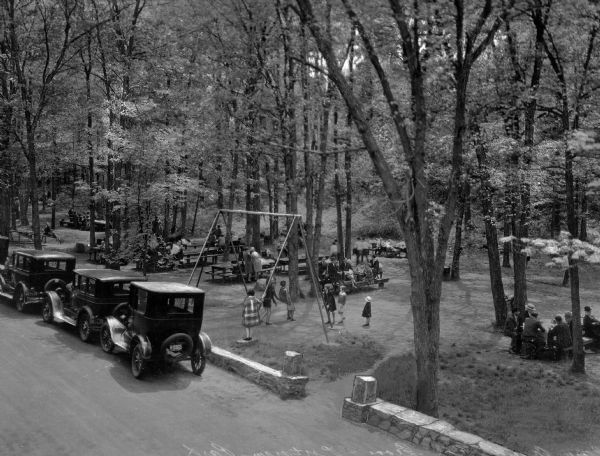 Elevated view across road towards Grove-Putnam Park. A short, stone wall separates automobiles parked on the road near the park. People are sitting at picnic tables and on benches while children play on the playground.