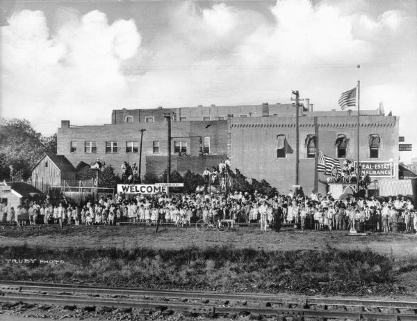 View taken from across railroad tracks of a children's park. Crowds of children and adults stand behind a fence along the park. Behind them are large brick buildings. A sign reads: "Welcome, Children's Park."