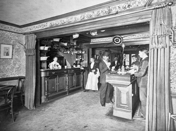 Interior of the bar room at Hotel Astor, built in 1904. Waiters, bartenders, and patrons stand around the bar, decorated in the Old World style.