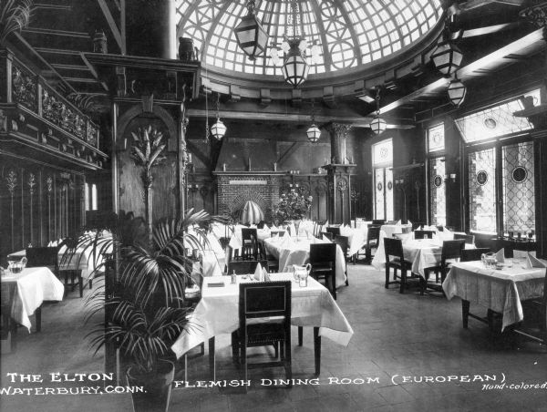 Interior of the Flemish Dining Room at the Elton Hotel, built in 1905. Decorative lighting surrounds a domed skylight, and below, tables are set for a meal. Large windows are on the right and a brick fireplace adorns the back wall. Caption reads: "Flemish Dining Room (European)."