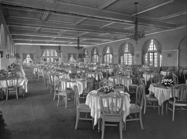 View of the dining room at Montauk Manor, which opened in 1927 and was designed by Carl Fisher. Large windows line the walls and tables are set with flower arrangements.