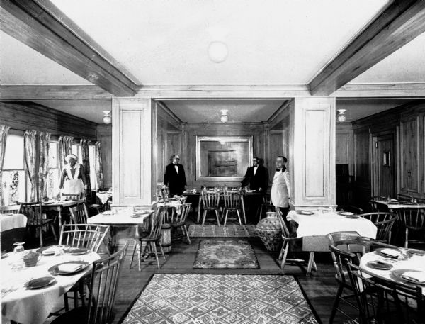 Interior of the dining room at the Terry Tavern. Waiters and a waitress stand near tables set for dining. Windows line the left wall and a door leads out of the dining room on the right.