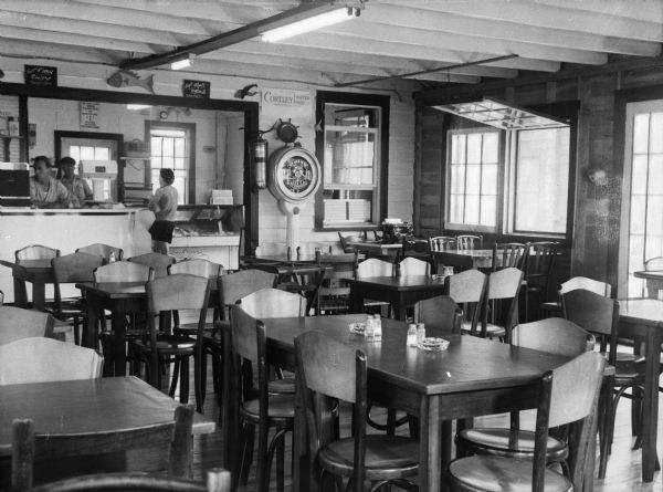 Interior of a sea food restaurant. Two men stand behind the counter where a woman waits. Chairs and tables crowd the room. Windows line the right wall and signs are behind the counter.