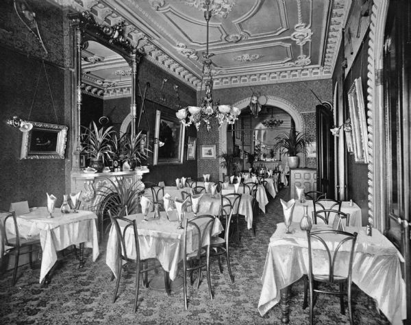 Interior of a restaurant at Hotel Astor, built in 1904. The room features chairs, tables, potted plants, and lavish decor. Along the walls are framed paintings and a mirror over a fireplace, and a decorative ceiling.