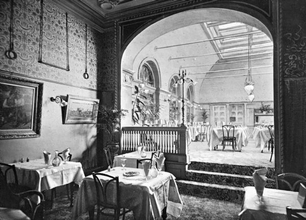 Interior of a restaurant at Hotel Astor, built in 1904. View from a room with tables and chairs toward a short staircase in an archway that leads to a room with more tables and chairs under a skylight.