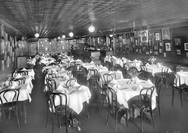 Interior of a restaurant and bar at American Art Studio. Tables and chairs fill the room and a bar is in the right corner. Framed art lines the walls.