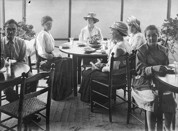 Several women and a man are sitting at tables while dining at a restaurant. Potted plants are in the background.